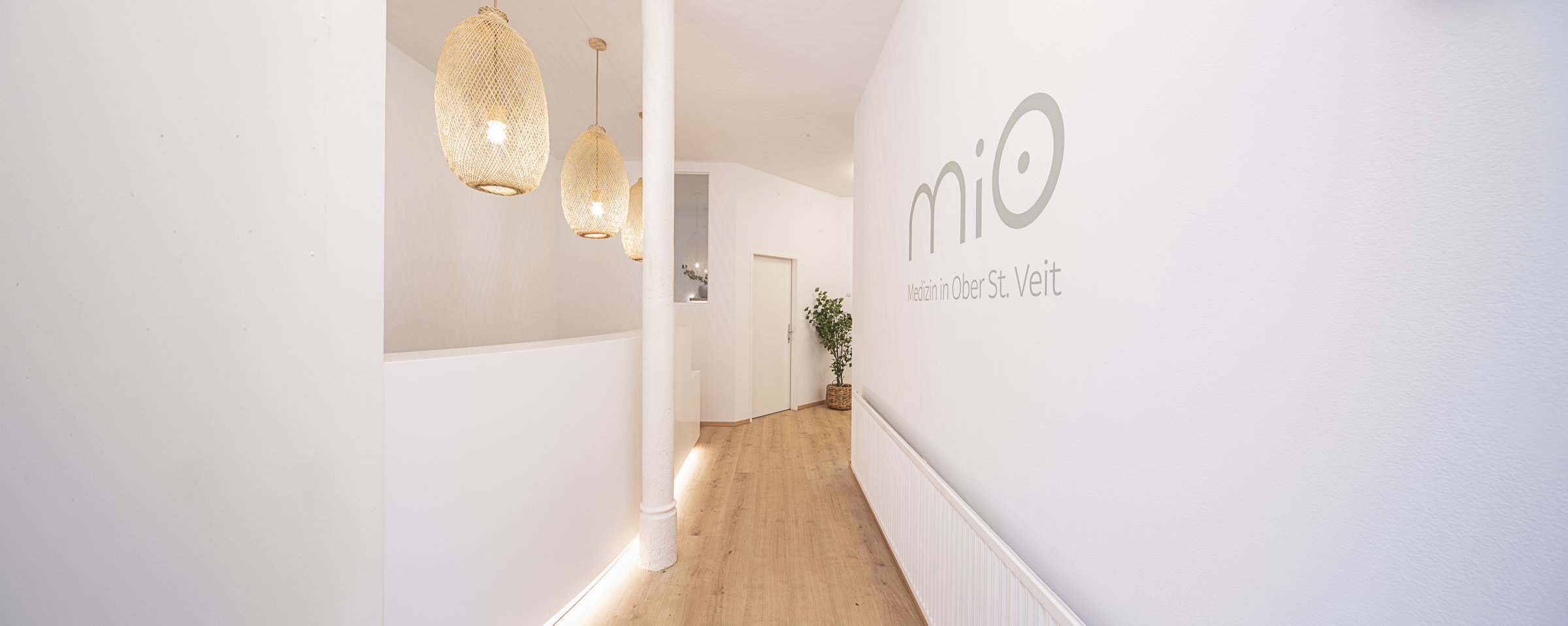 Empfangsbereich im MIO Medizin Wahlarztzentrum Ober St. Veit – mit Wandlogo, Korbleuchten und warmem Holzboden